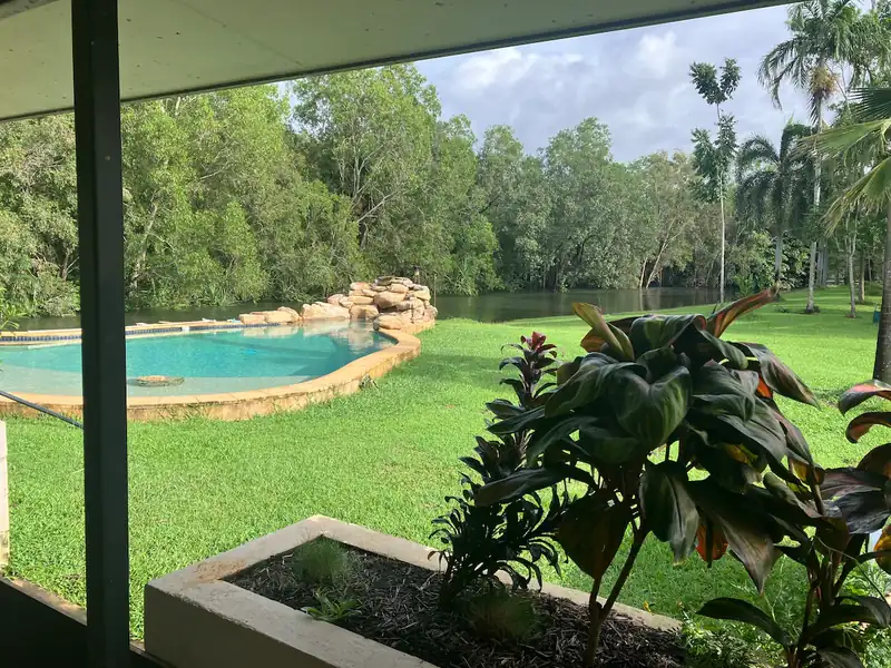 Green lawn and a stone-edged pool beside a wooded waterway, seen from a shaded terrace